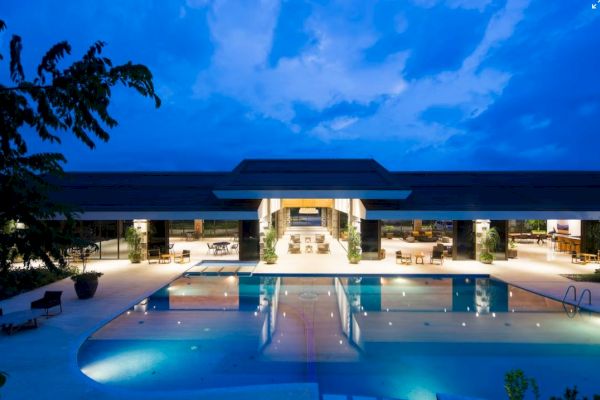 A luxurious, modern building with large glass doors is reflected in a tranquil swimming pool under a dramatic blue evening sky.