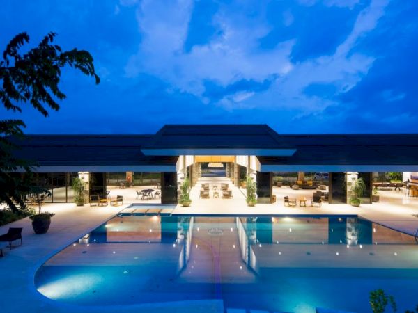 A luxurious, modern building with large glass doors is reflected in a tranquil swimming pool under a dramatic blue evening sky.