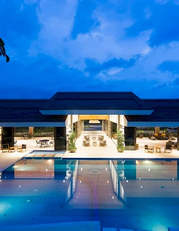 The image shows a luxurious building with a large, illuminated swimming pool in the foreground, under a twilight sky.
