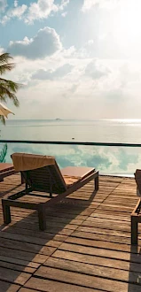 Poolside scene with lounge chairs, umbrellas, palm trees, and a view of the ocean under a cloudy sky.