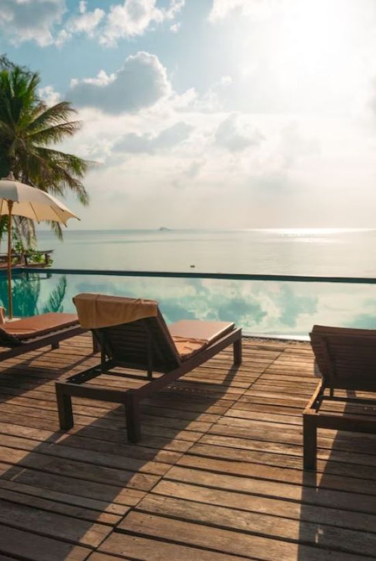 A serene poolside scene with sun loungers, umbrellas, and palm trees overlooking a calm body of water under a partly cloudy sky.
