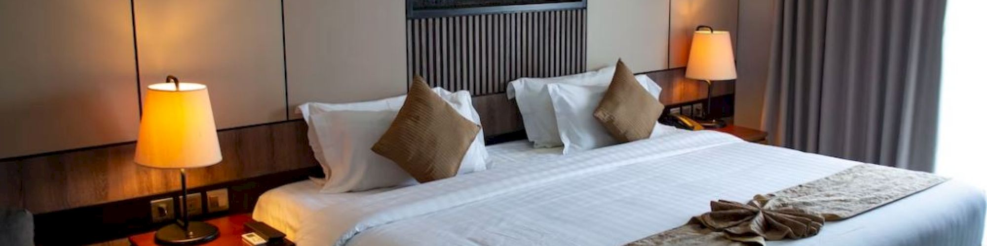 A cozy hotel room with a large bed, two bedside tables with lamps, a decorative wall feature, and a window with curtains.