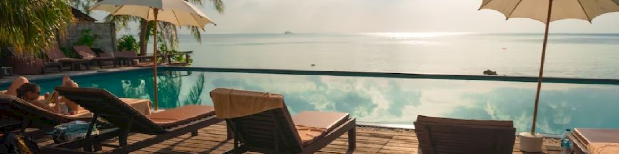 A serene poolside scene with lounge chairs, umbrellas, and palm trees, overlooking a calm sea under a partially cloudy sky.