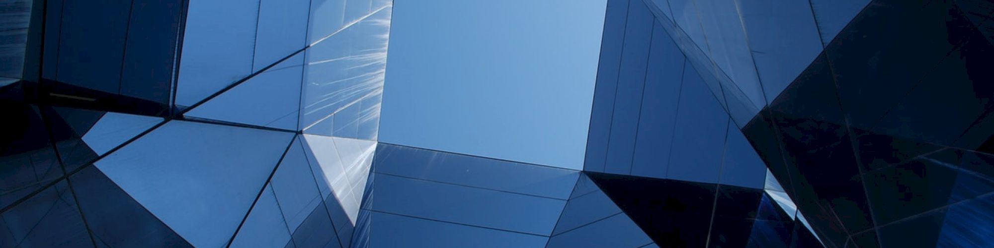 The image shows an abstract view of a modern building with reflective glass surfaces forming geometric shapes against the sky.
