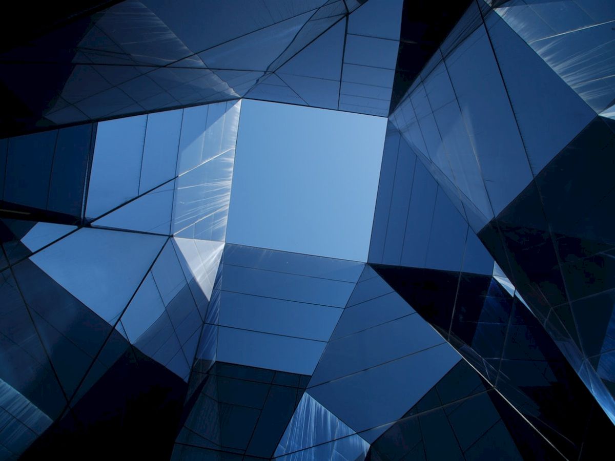 Geometric glass structures forming a symmetrical pattern frame a view of the blue sky above.