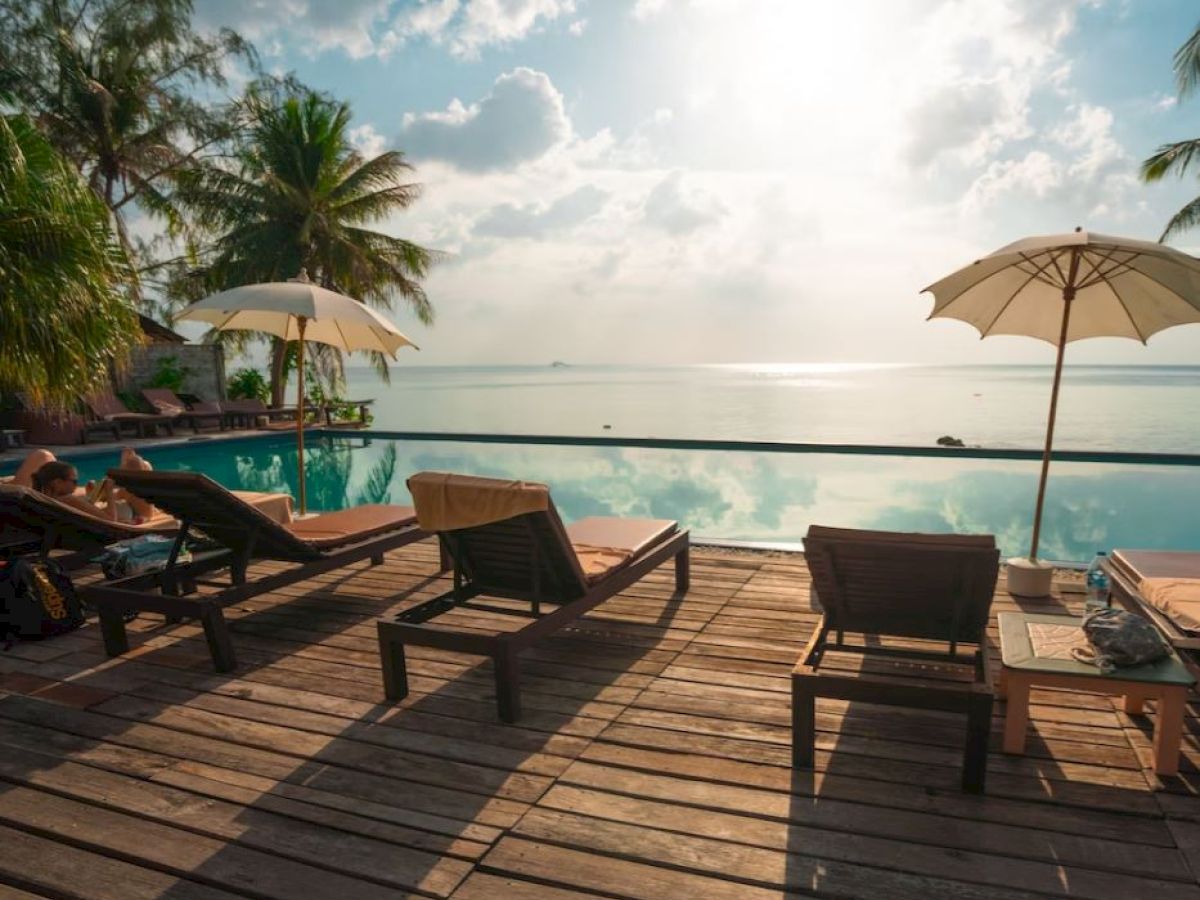A serene poolside scene with lounge chairs, umbrellas, and a view of the ocean under a partly cloudy sky.