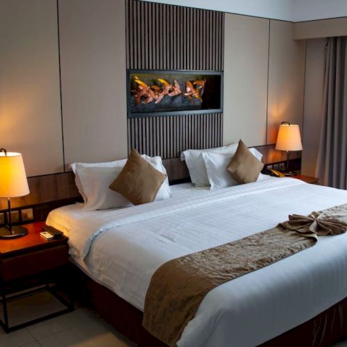 A cozy hotel room featuring a large bed with decorative pillows, bedside lamps, and a wall-mounted art piece, creating a warm ambiance.