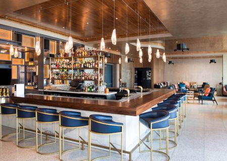 A modern bar with blue stools, wooden accents, hanging lights, and a fully stocked back bar. Lounge seating is visible in the background.