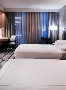 A modern hotel room with two beds, a wall-mounted TV, desk, chair, and stylish decor. It features large windows with curtains.