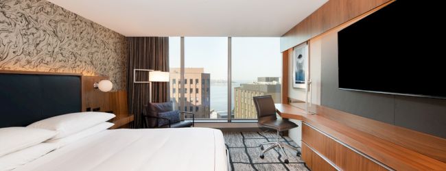 A modern hotel room features a bed, desk, large TV, and a window with an urban view, creating a relaxing environment.