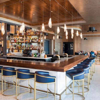 A modern bar with a wooden counter, blue chairs, pendant lights, and a well-stocked liquor shelf, set in a stylish lounge area.