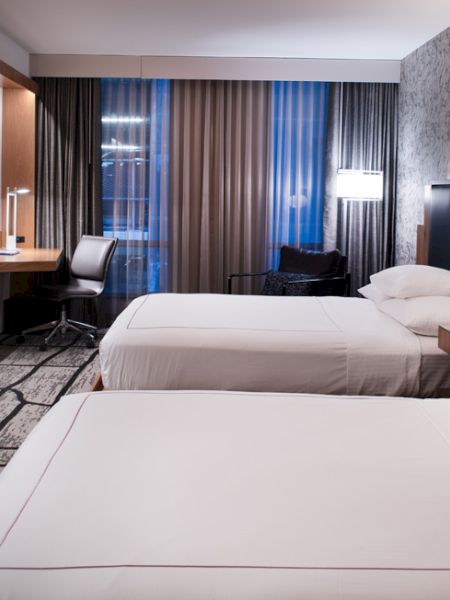 The image shows a modern hotel room with two neatly made beds, a desk with a chair, a flat-screen TV, and contemporary lighting.