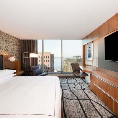 A modern hotel room with a large bed, wall-mounted TV, desk, and window view of city buildings and water.