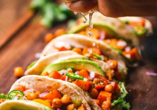 A hand squeezes lime over tacos filled with chickpeas, avocado slices, and vegetables on a wooden board. Fresh and colorful.