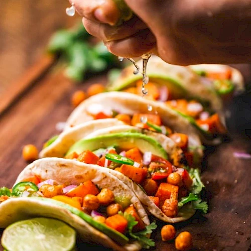 A hand squeezes lime over tacos filled with chickpeas, avocado slices, and vegetables on a wooden board. Fresh and colorful.