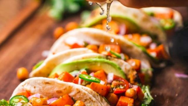A hand squeezes lime over tacos filled with chickpeas, avocado slices, and vegetables on a wooden board. Fresh and colorful.