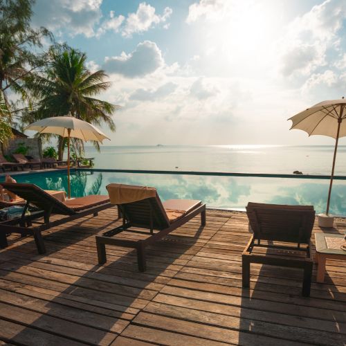 Lounge chairs and umbrellas by a pool, facing the ocean with palm trees and a tranquil sky in the background.