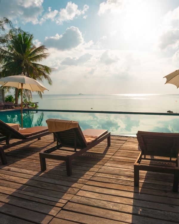 Lounge chairs and umbrellas by a pool, facing the ocean with palm trees and a tranquil sky in the background.