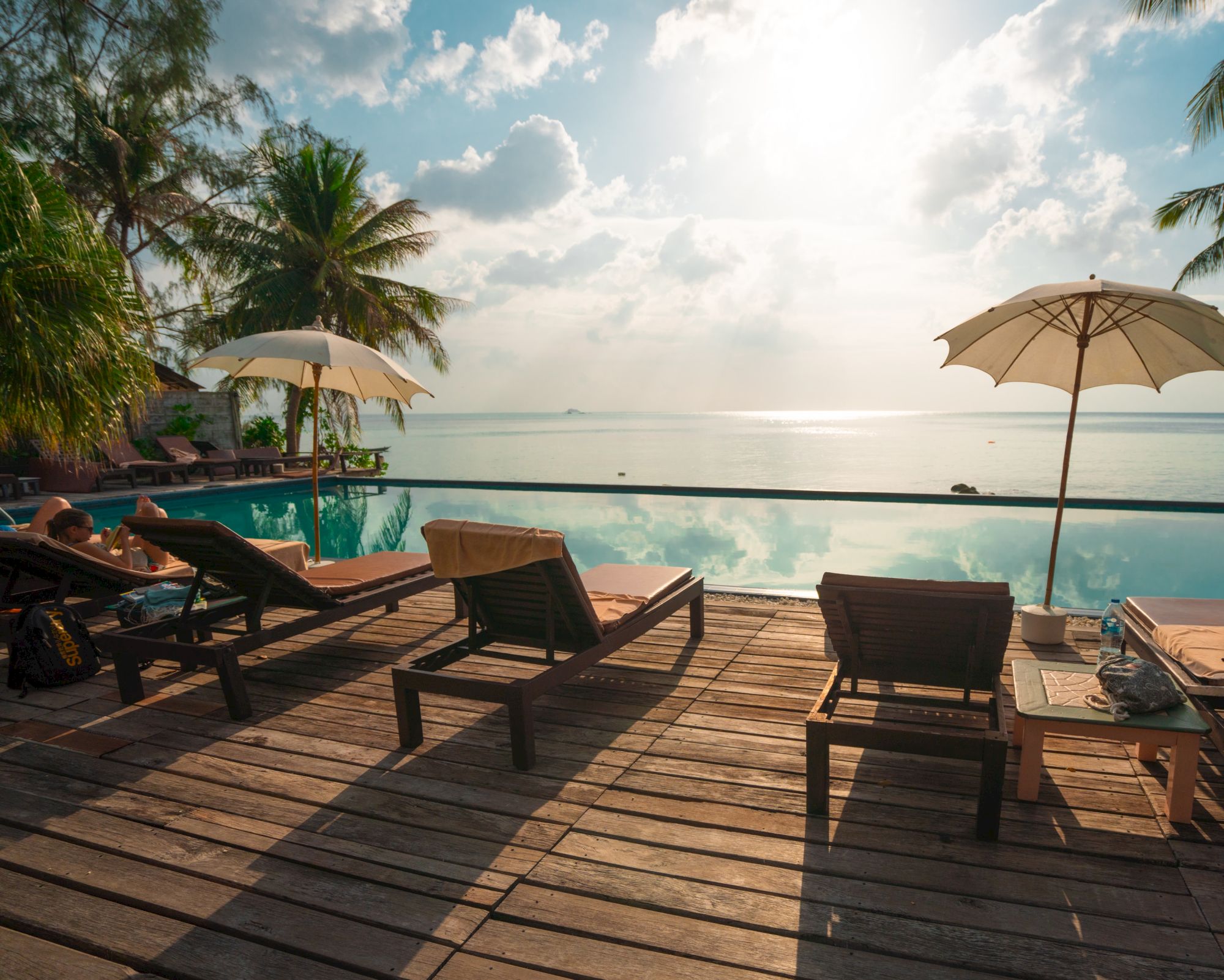 Poolside with lounge chairs, umbrellas, overlooking a calm sea at sunrise, surrounded by palm trees and clear skies.