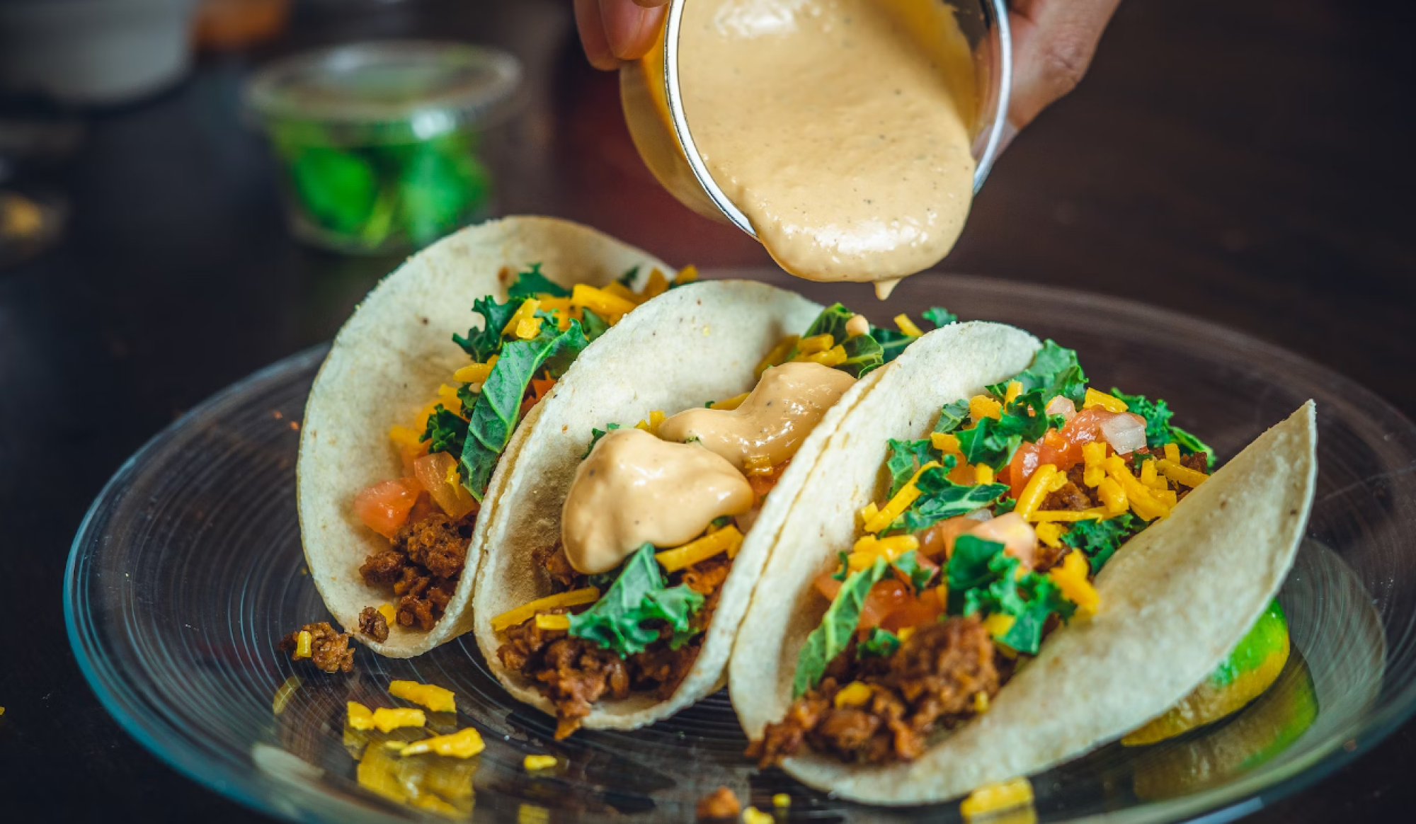 Three tacos on a plate with toppings like cheese, tomatoes, greens, and sauce being poured over them.