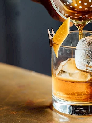 A hand pours a brown liquid into a glass with ice and a citrus peel garnish, set on a wooden surface.