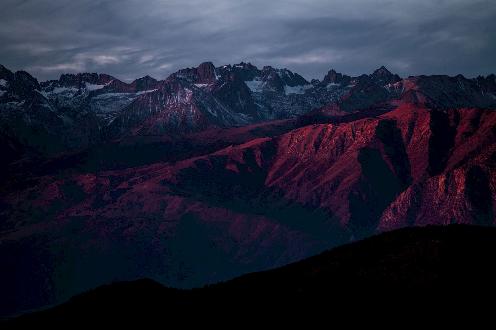 A mountain range under a dramatic sky, with peaks and slopes illuminated by red and purple hues.