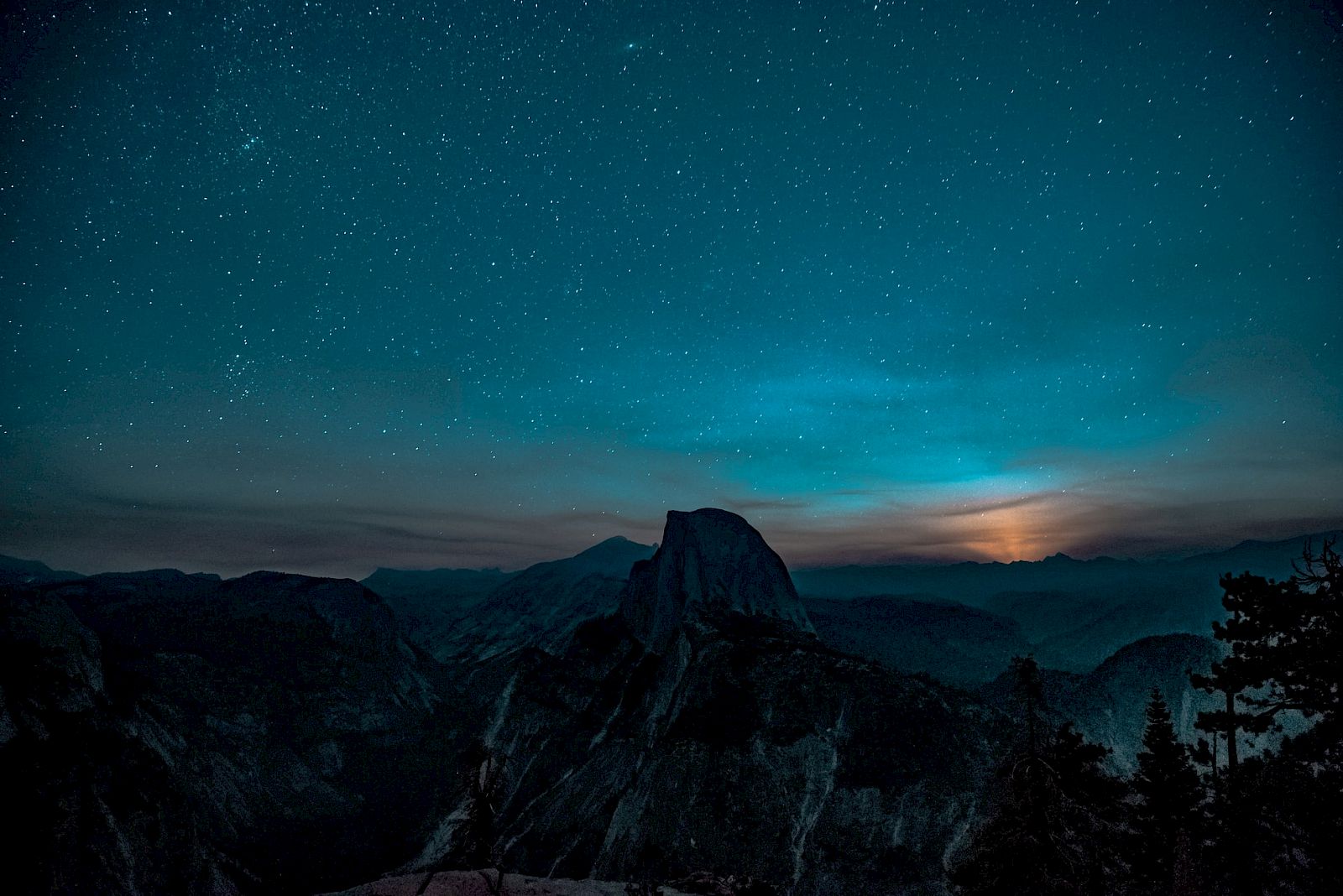 A starry night sky over a mountain landscape with a glowing horizon and silhouetted trees in the foreground.