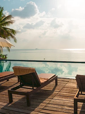 Sunlit poolside with lounge chairs, umbrellas, palm trees, and an ocean view under a blue sky.