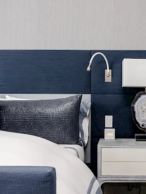 A modern bedroom with a navy headboard, white bedding, gray nightstand, decorative pillows, a lamp, and a wall light.
