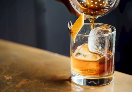 A cocktail being poured into a glass with large ice cubes and a lemon twist garnish on a countertop.