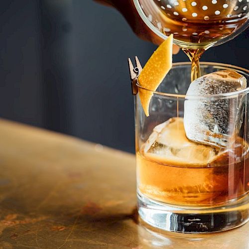 A cocktail being poured into a glass with large ice cubes and a lemon twist garnish on a countertop.