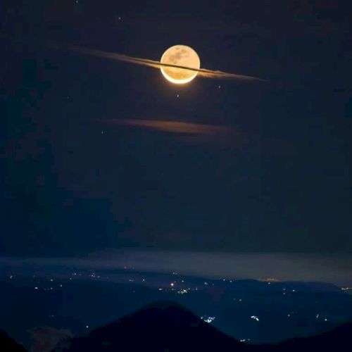 A view of the moon with horizontal clouds, resembling Saturn’s rings, above a landscape with scattered lights in the distance.