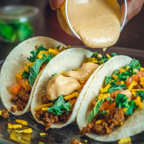 Three tacos with ground meat, cheese, lettuce, and sauce being poured on top.