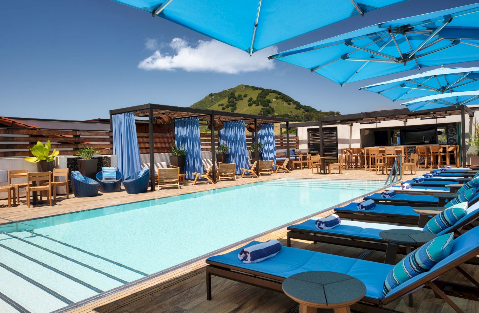 Rooftop pool with lounge chairs, cabanas, and blue umbrellas under a sunny sky. Hills visible in the background create a serene setting.