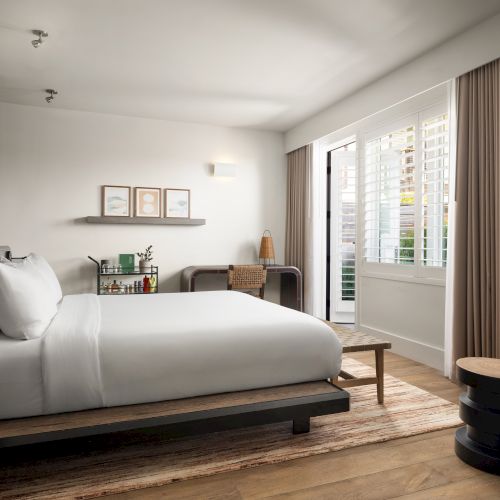 Bright bedroom with a modern design, featuring a bed, chair, desk, and art on the wall. Natural light filters through the curtains.
