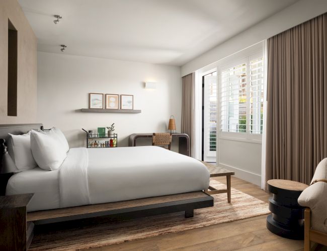 Bright bedroom with a modern design, featuring a bed, chair, desk, and art on the wall. Natural light filters through the curtains.