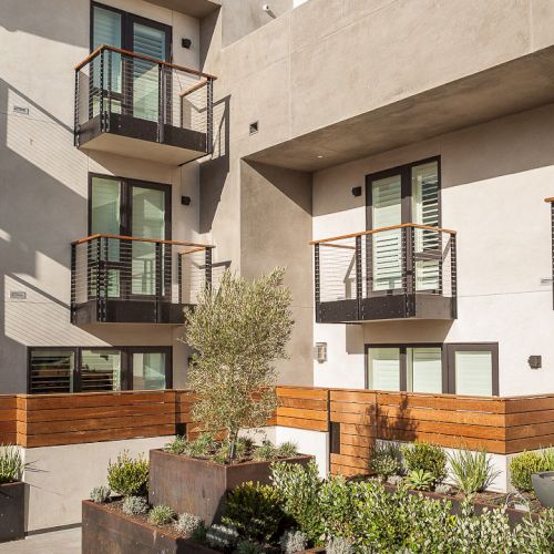 A modern apartment building with balconies and a courtyard featuring planters filled with green plants and shrubs.