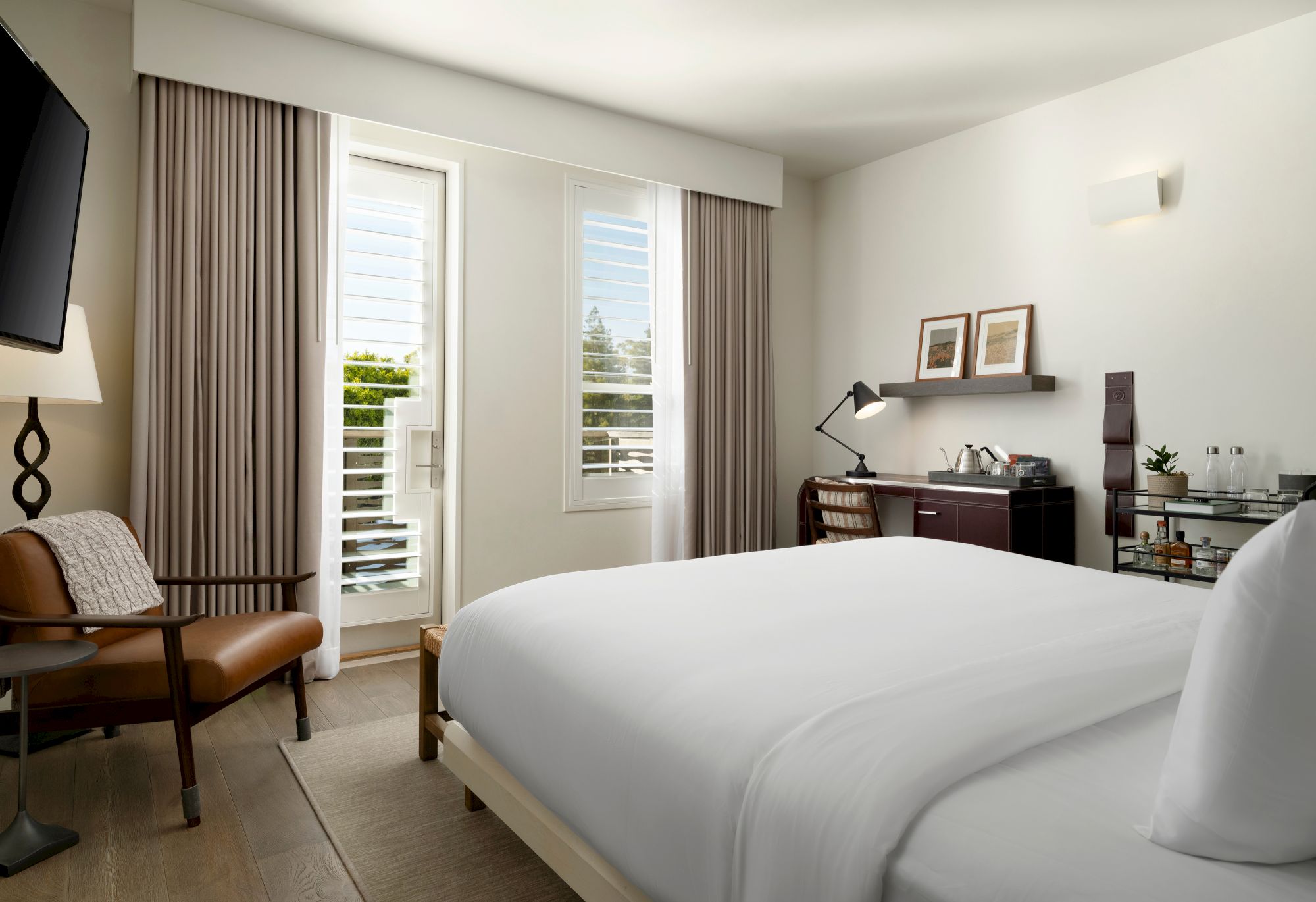 A modern hotel room with a large bed, desk, chair, TV, and elegant decor, featuring a window with shutters to let in natural light.