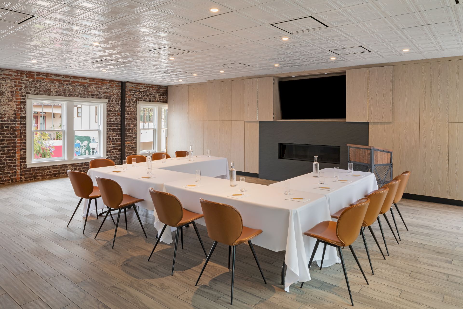 A modern meeting room with a U-shaped table setup, brown chairs, a TV, and large windows, featuring wooden and brick walls, is shown.