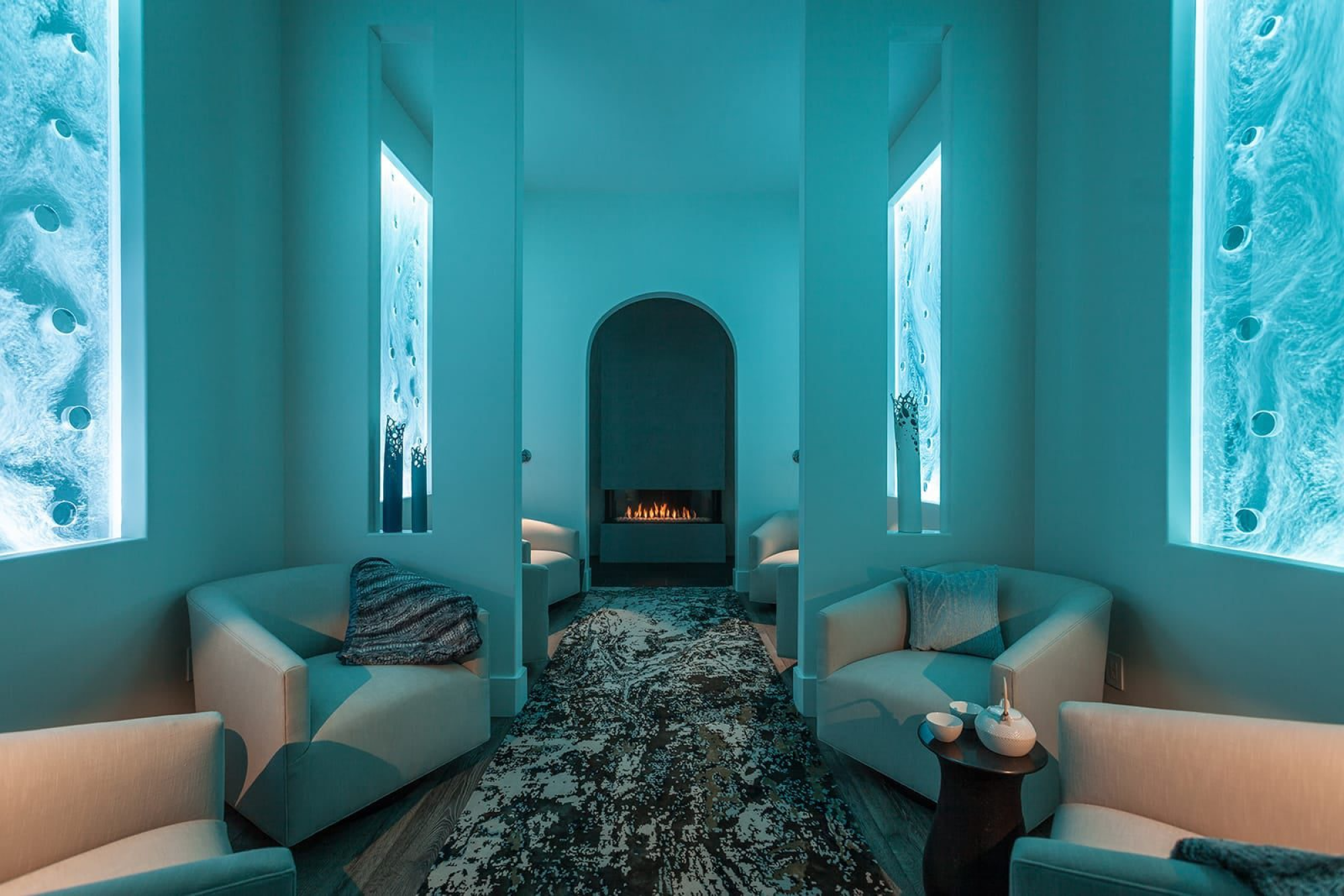 A modern lounge with blue lighting features armchairs, a fireplace, abstract wall art, and a patterned rug.