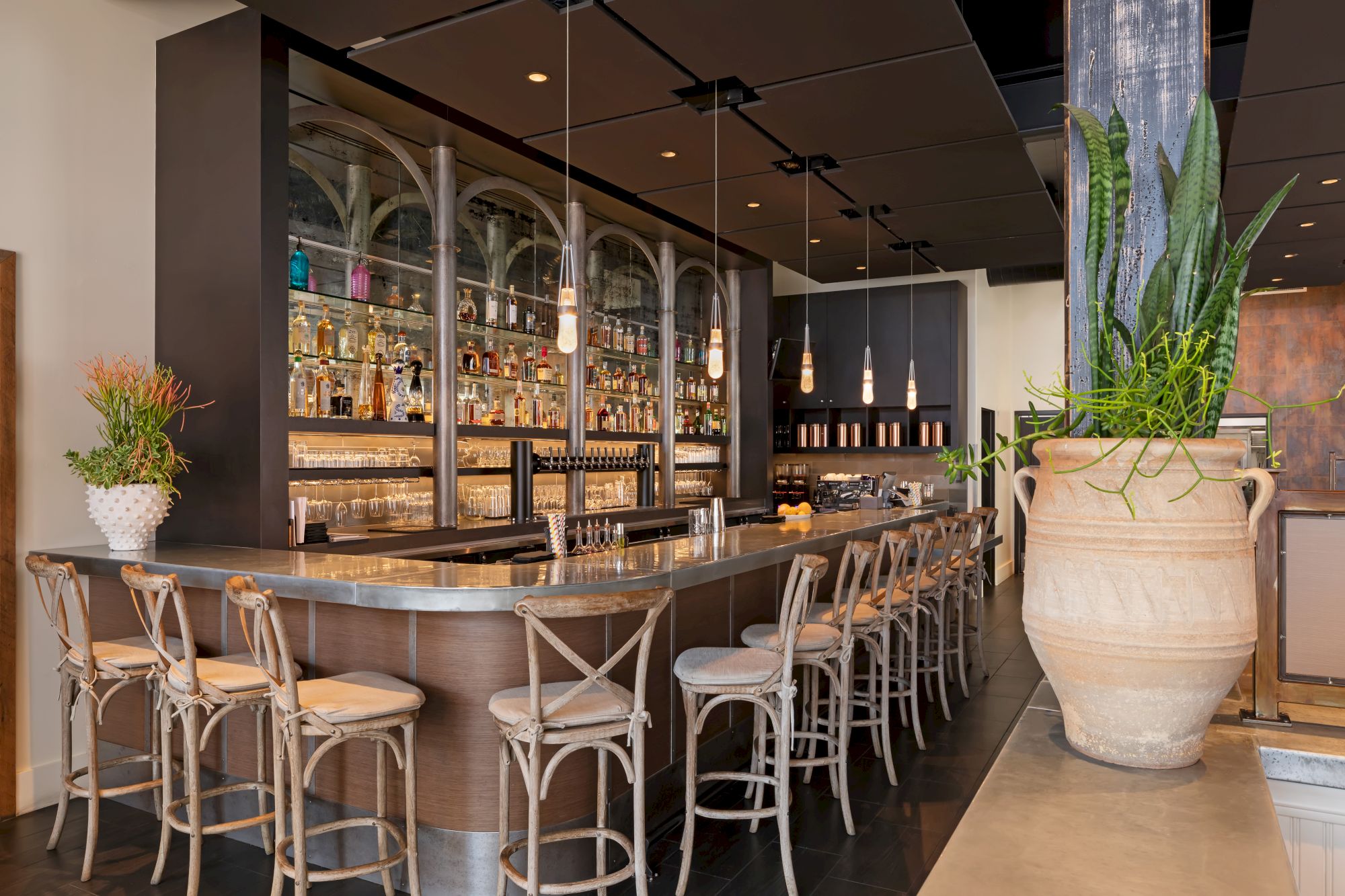 The image shows a stylish bar with wooden chairs, a sleek counter, hanging lights, and a well-stocked liquor shelf.