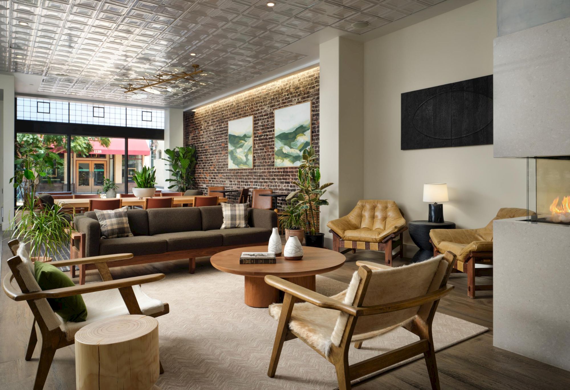 A modern living room with wooden furniture, plants, a fireplace, and artwork on brick and white walls, creating a cozy atmosphere.