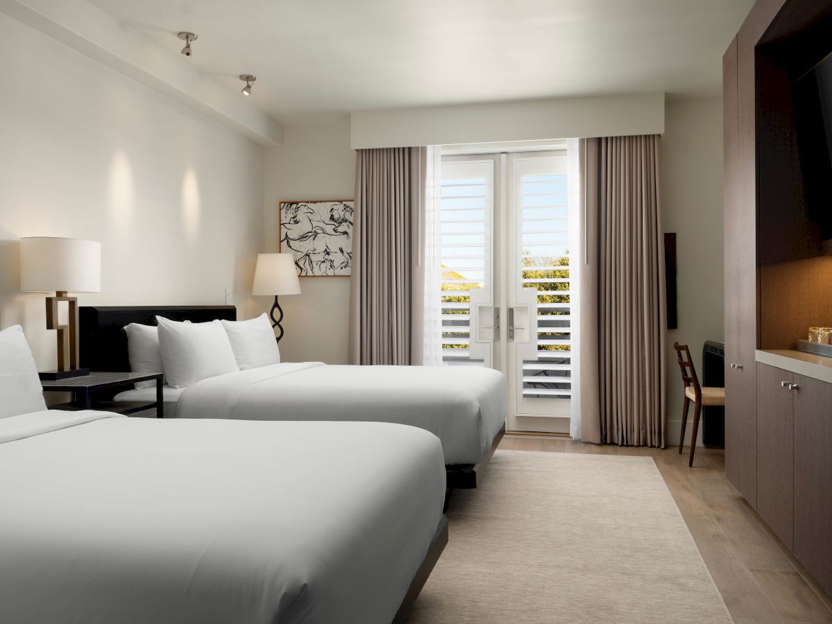 A modern hotel room with two beds, a nightstand, lamps, artwork, a desk, a TV, and a view through shuttered doors.