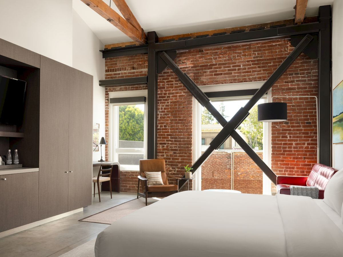 A modern room with exposed brick, a bed, desk, chair, TV, and large windows. There are beams, a red sofa, and a greenery view outside.
