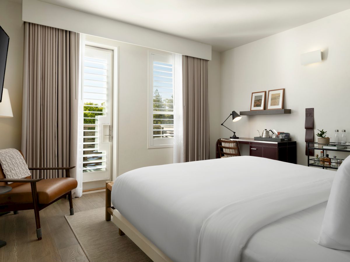 A modern hotel room with a bed, desk, chair, and TV. The room has a window with shutters and various decor items on shelves.