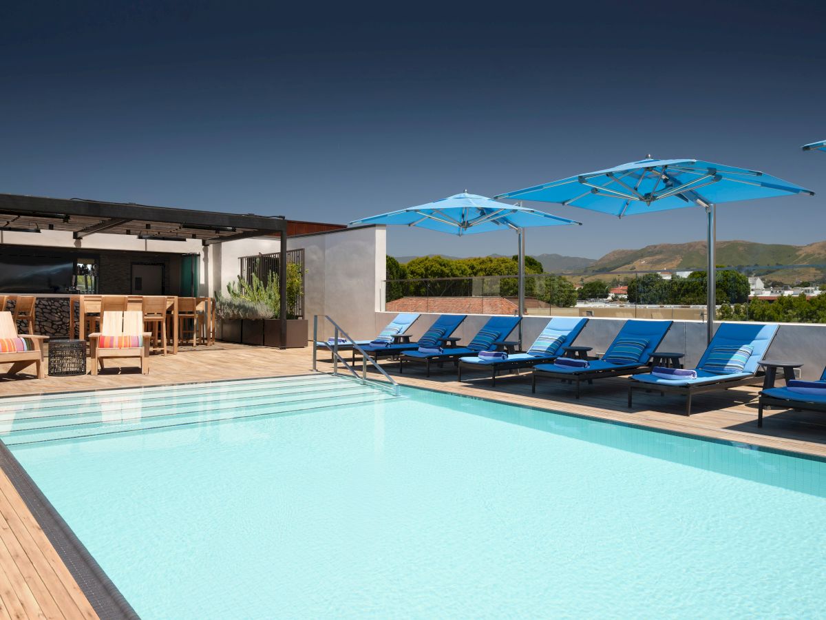 A poolside area with loungers, blue umbrellas, and a bar, set against a backdrop of mountains under a clear sky.
