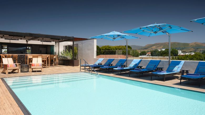 A poolside area with loungers, blue umbrellas, and a bar, set against a backdrop of mountains under a clear sky.