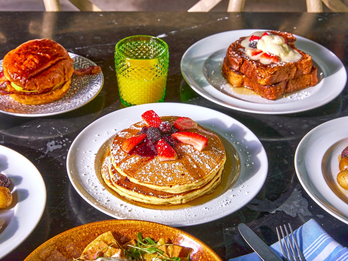 A breakfast spread with pancakes topped with berries, French toast, a sandwich, roasted potatoes, and a glass of orange juice on a table.
