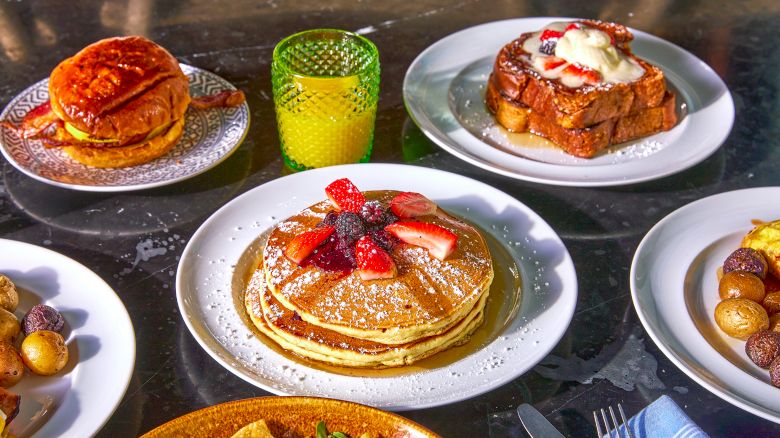 A breakfast spread with pancakes topped with berries, French toast, a sandwich, roasted potatoes, and a glass of orange juice on a table.