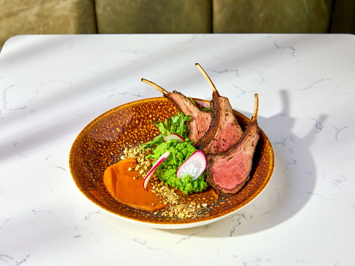 A plate with sliced lamb chops, green puree, radish slices, crushed nuts, and a sauce, set on a marbled table.